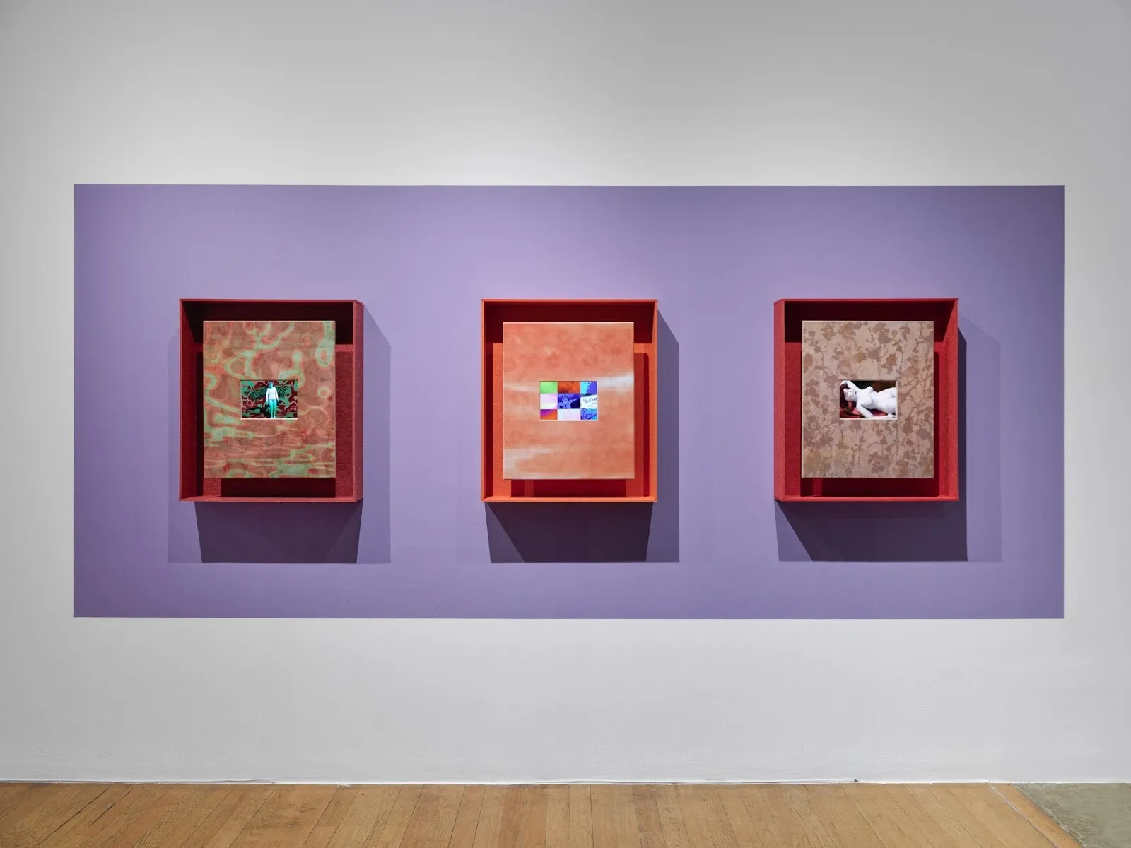 A wall is painted white with an interior purple rectangle. Within the rectangle, three similarly-sized paintings hang in a row, each in a boxy red or orange frame. Each painting comprises of a patterned exterior which houses a smaller interior image.