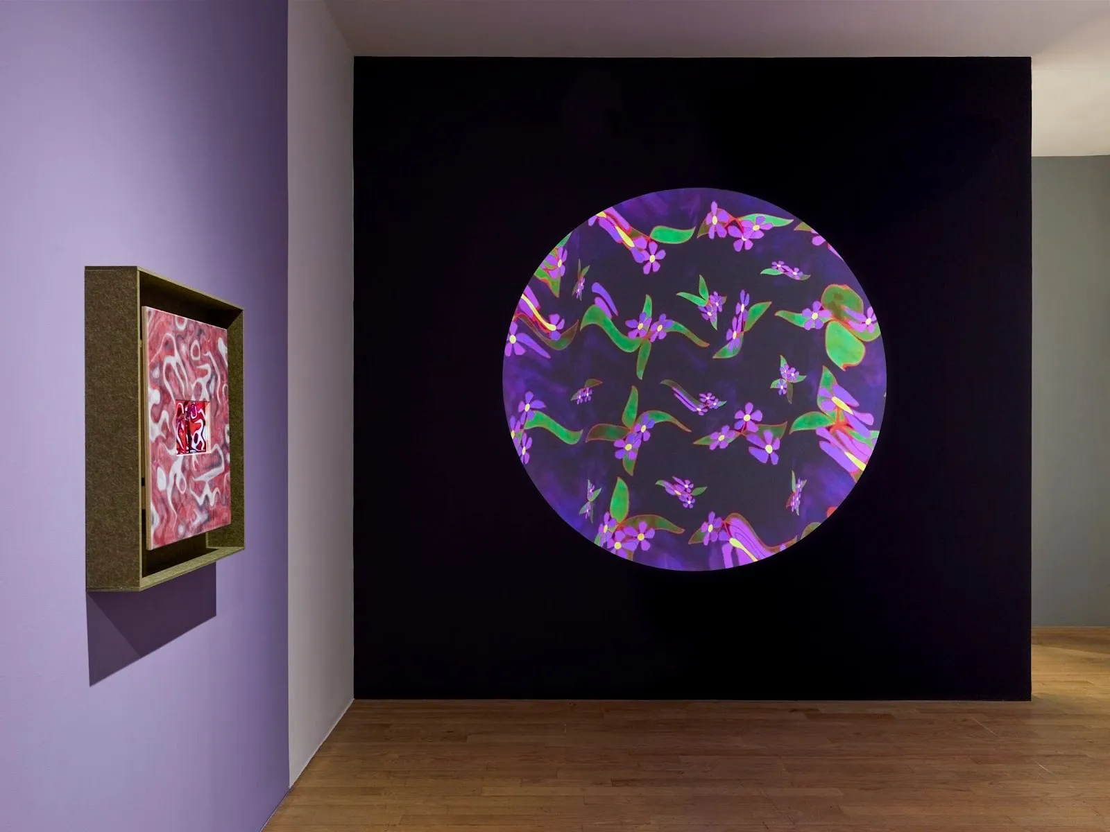 Purple and white-painted wall and a black painted wall meet. On the purple wall, a green-framed pink and white painting hangs. On the black wall, a round projection displays purple and green floral patterns.