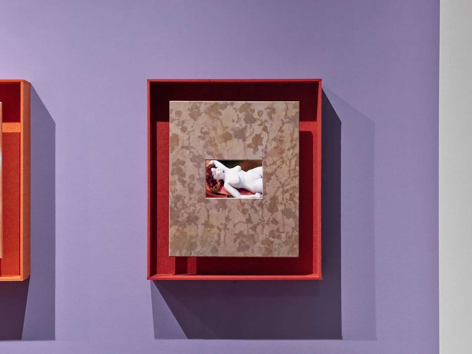 A purple wall features a red-framed painting. The painting has a patterned exterior, which frames a smaller central image of a nude woman figure in repose against a red background.