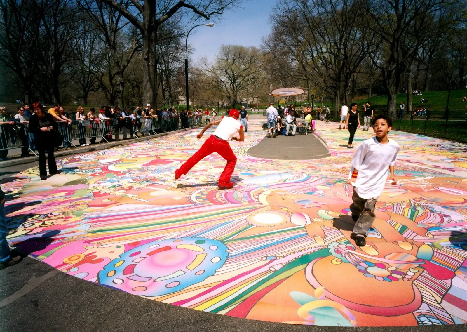In a park, a skate rink floor is decorated with colorful patterns and illustrations. People and children skate and walk on the surface, which is barricaded as people watch from the sidelines.