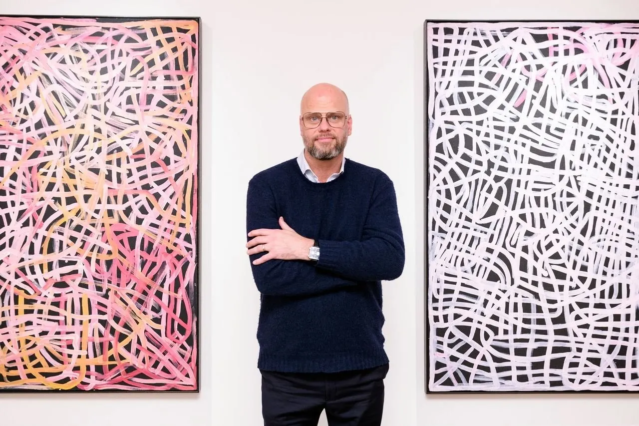 A man in a blue sweater with glasses and a beard stands in between two paintings of the same size and style. The paintings feature curling lines that intersect, almost like a street map. One painting has a pink and yellow palette, the other is white.