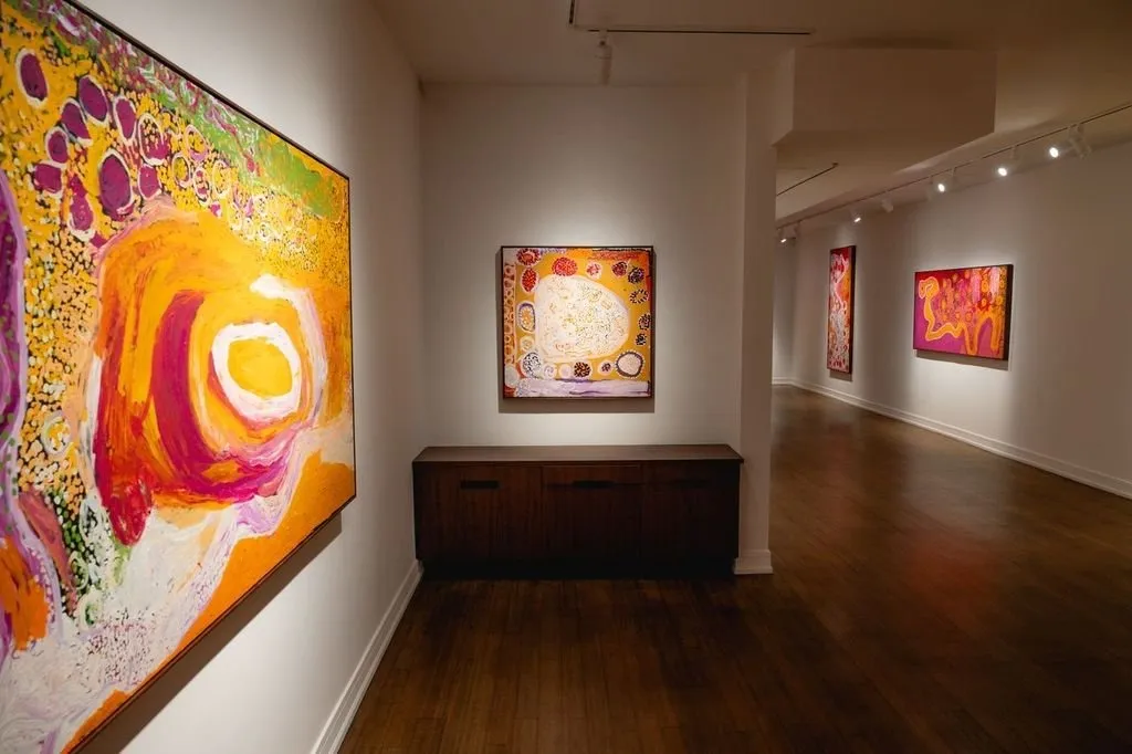 A white gallery room with dark wood floors features spotlit paintings on its walls. The paintings have vibrant, warm hues of orange, red, white, purple, and green, and abstract gestures and shapes.