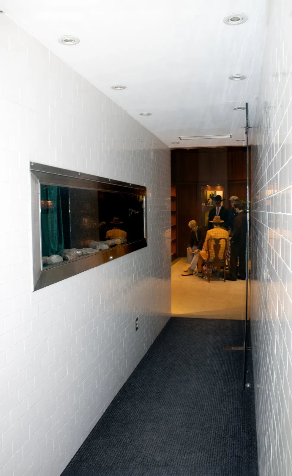 A white tiled hallway with grey carpeting leads into another room. A glass window is built into the side of the wall, where various bone-like objects against green velvet are displayed. In the back room, a staged set with mannequins can be seen.