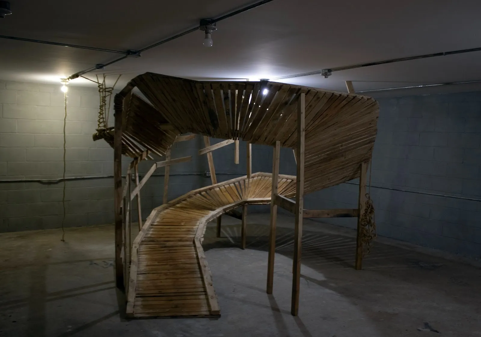 A dimly lit basement with concrete walls and flooring houses a large, wooden sculpture. The sculpture is a curving, scaffolded stairway/ramp, which spirals up and almost touches the ceiling.