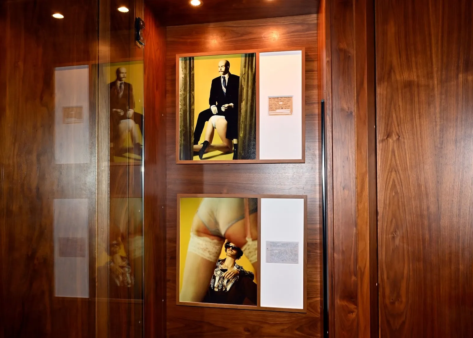 A wood paneled wall features two works of art. The framed works appear as paintings with semi-nude figures and men in suits, accompanied by a small block of text text on each image's right hand side.