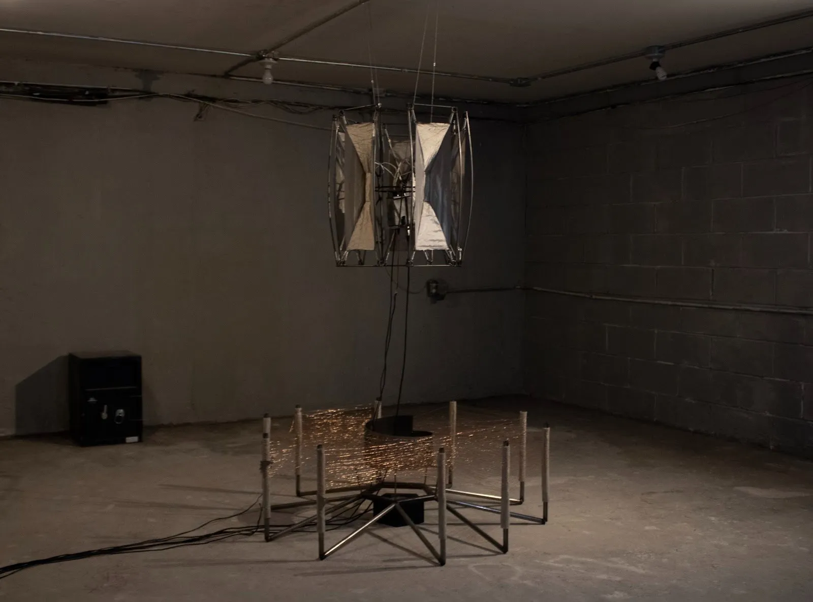 A multi-part sculpture in a dark room. The hanging piece appears as a bare speaker-antenna-like structure: flared panels arranged in a circle, facing out. Under, the floor piece is made of multiple bent pipes facing up with copper wires woven between
