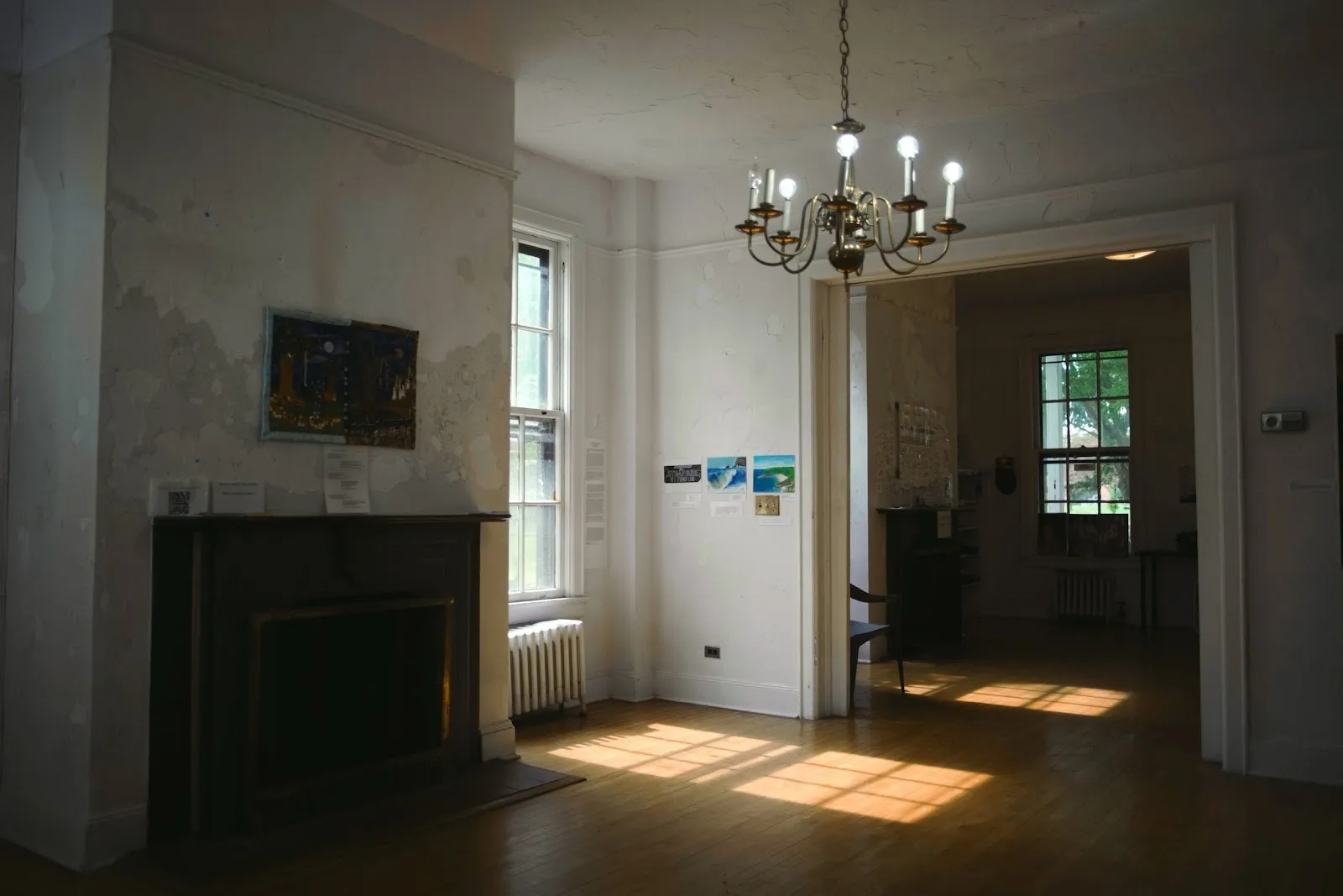 The interior of a house has wooden floors and peeling white walls. A small metal chandelier hangs, and multiple 2D works populate the walls. Light flows in from the windows.