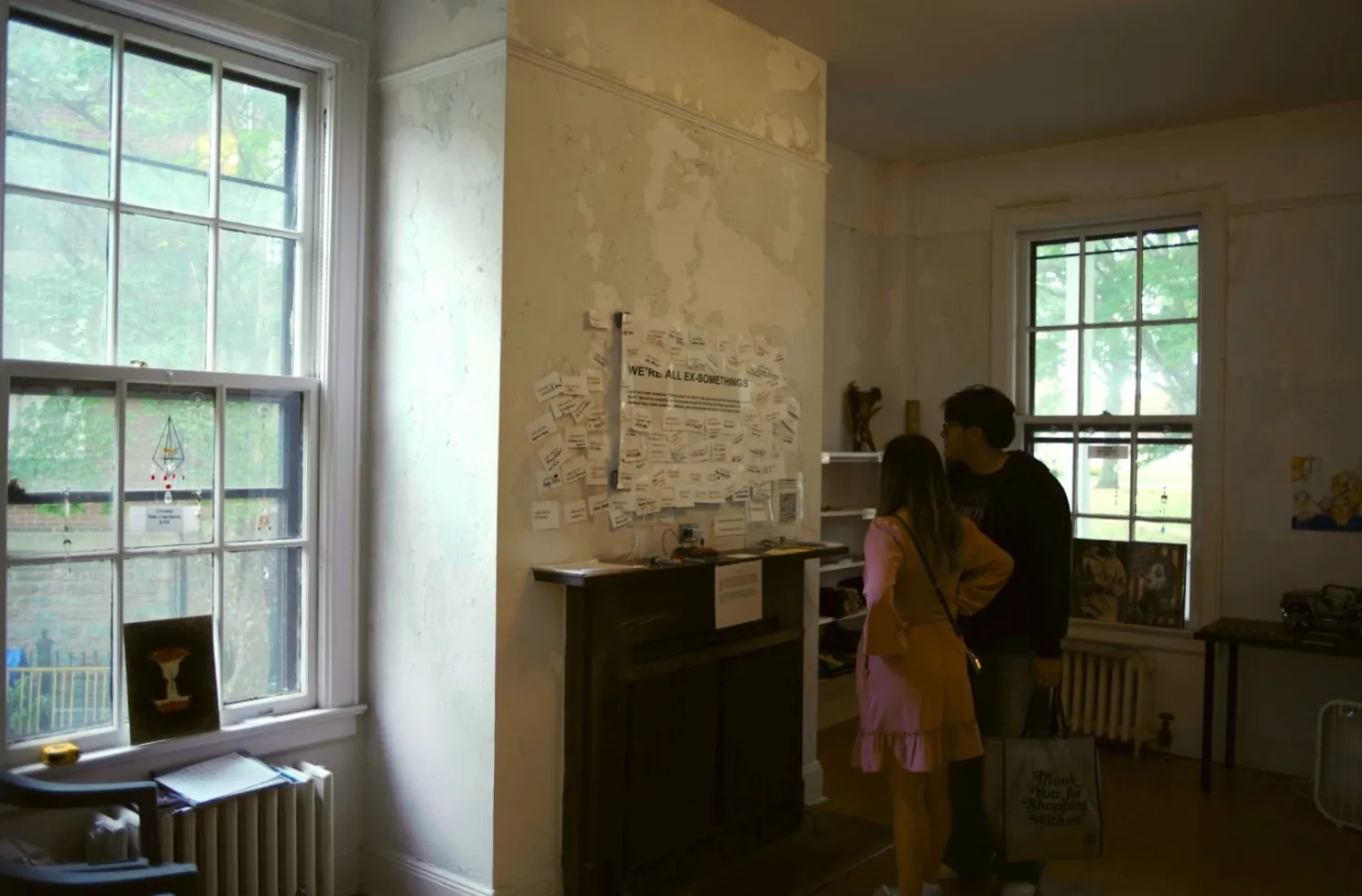 In a house with peeling white paint and large windows, people look at 2D works affixed to the walls.