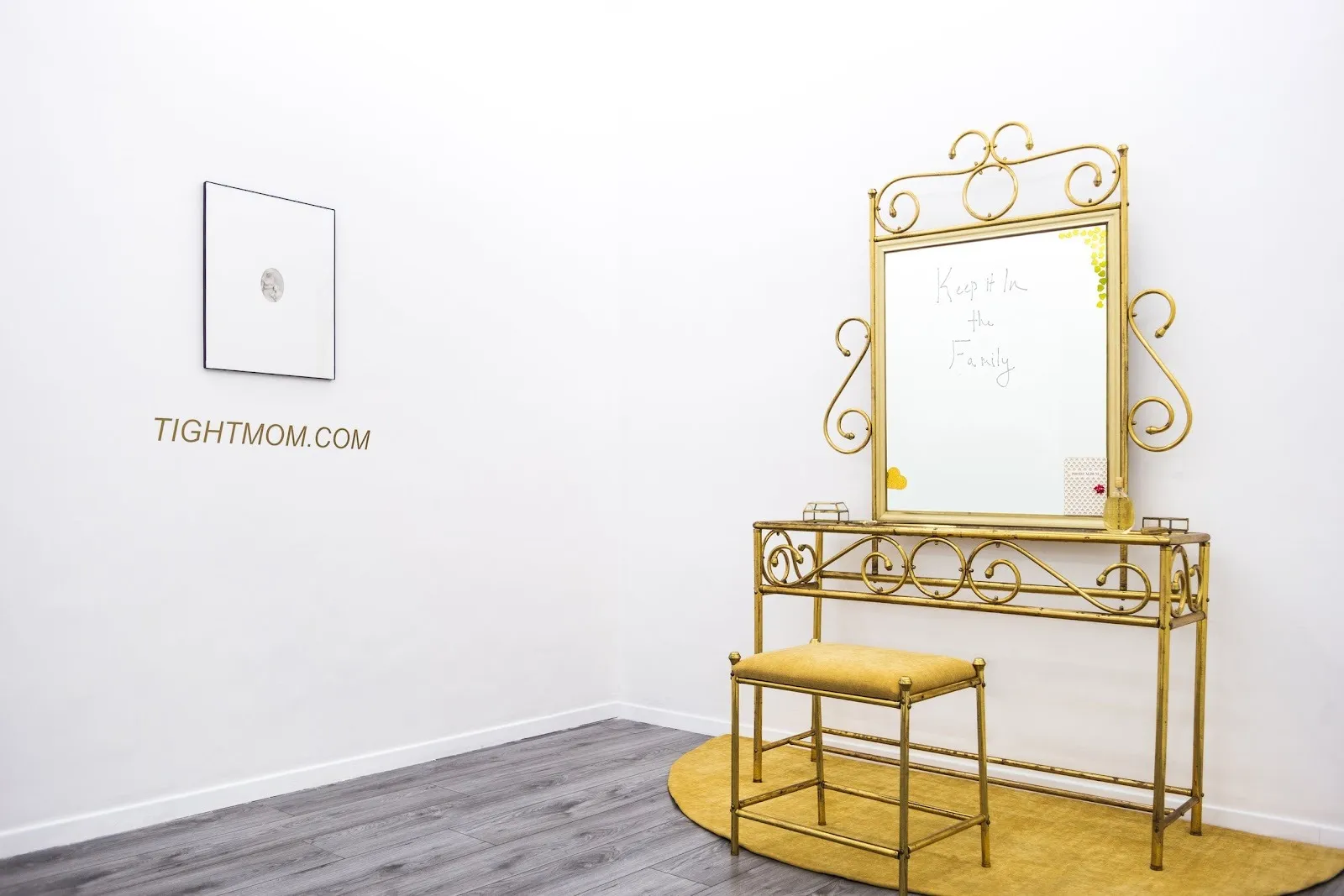 A room with white walls meets at the corner and features one wall work and one sculpture. The sculpture is a vanity table and stool, with writing on the mirror. The wall piece is a small, circular image, framed and hanging above wall text vinyl.