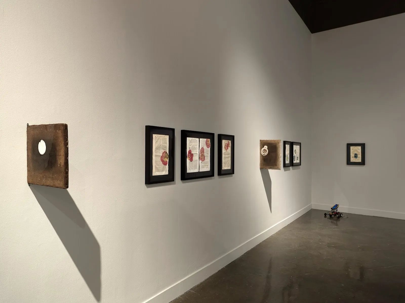 A white gallery wall features a variety of smaller wall-mounted works: framed ephemera and wooden boards that jut out perpendicular to the wall.