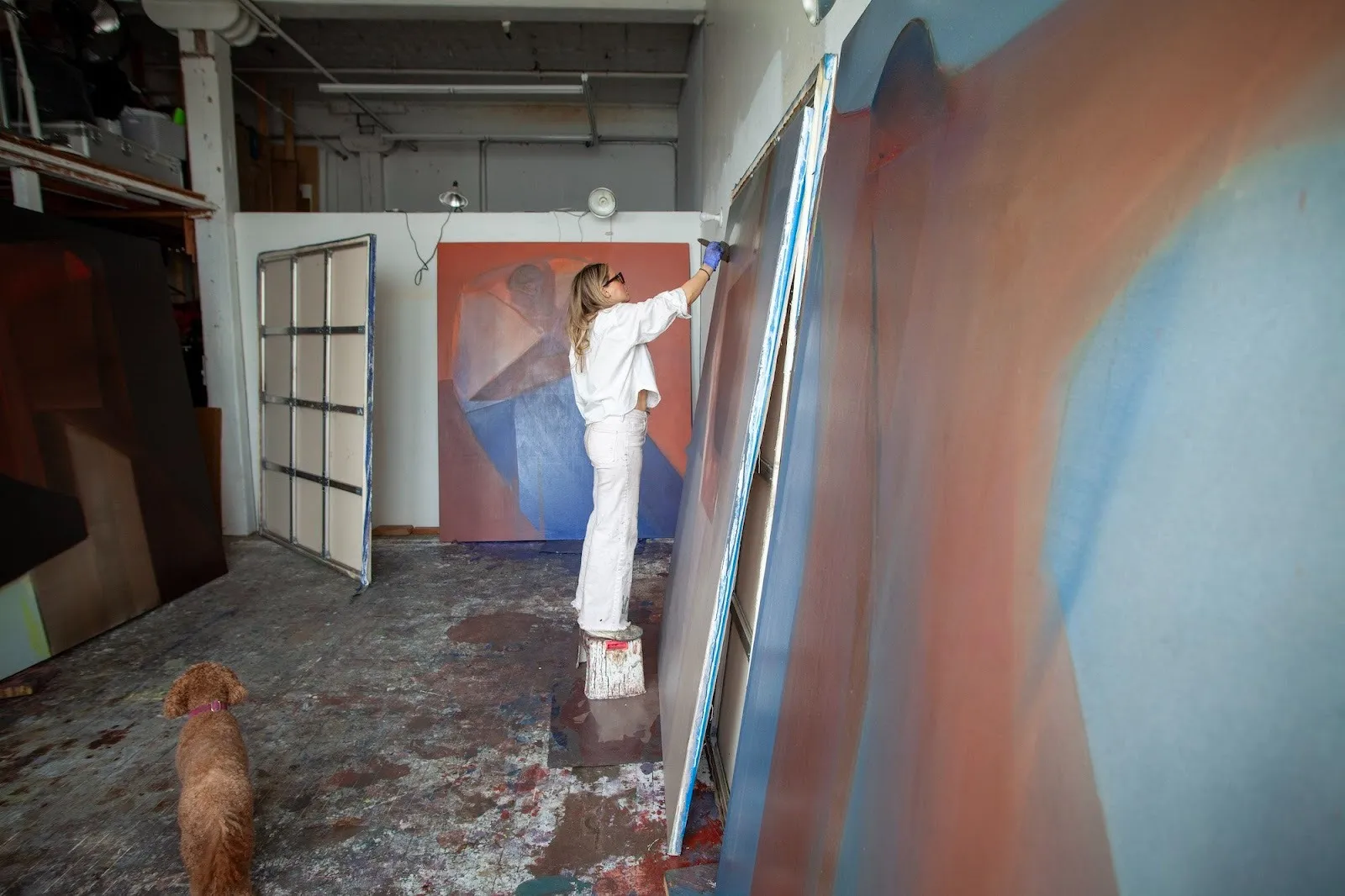 Artist Heidi Hahn wearing a white suit painting abstract color fields in her studio with a dog on the left side of the picture.