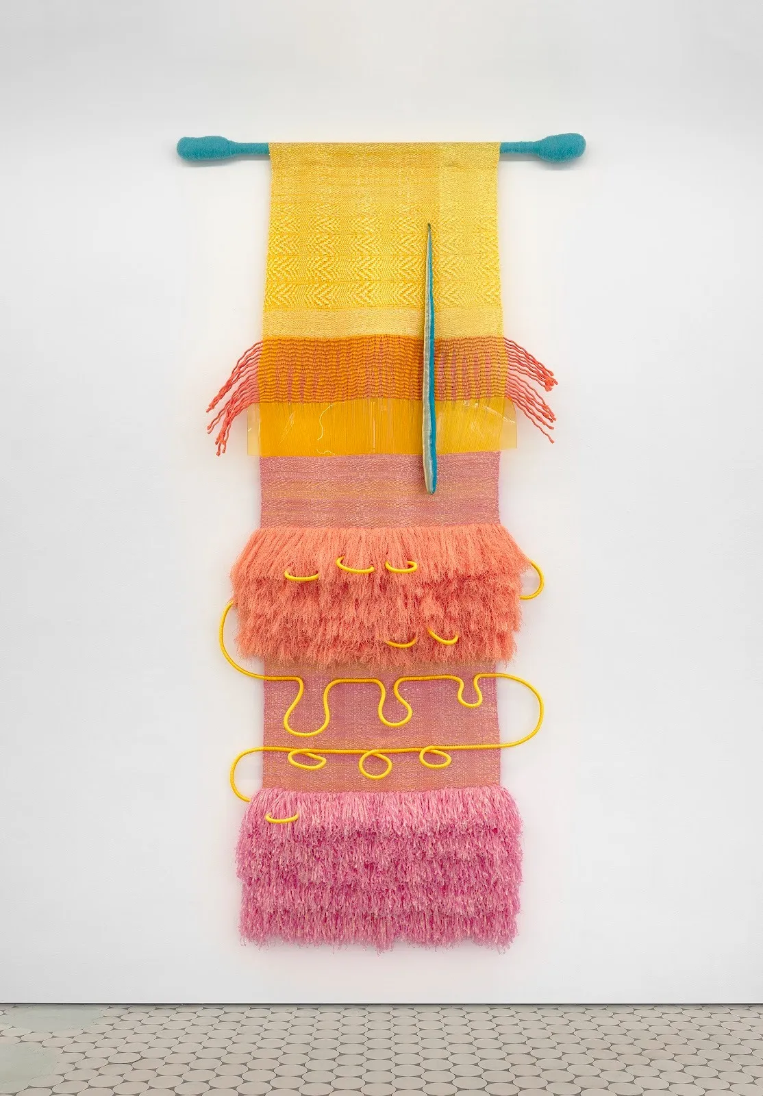 A fiber work mounted to a white wall is made from layers of yellow, pink, and orange fiber, some tufted, some thick and wound. The piece hangs from a rod wrapped with blue thread.
