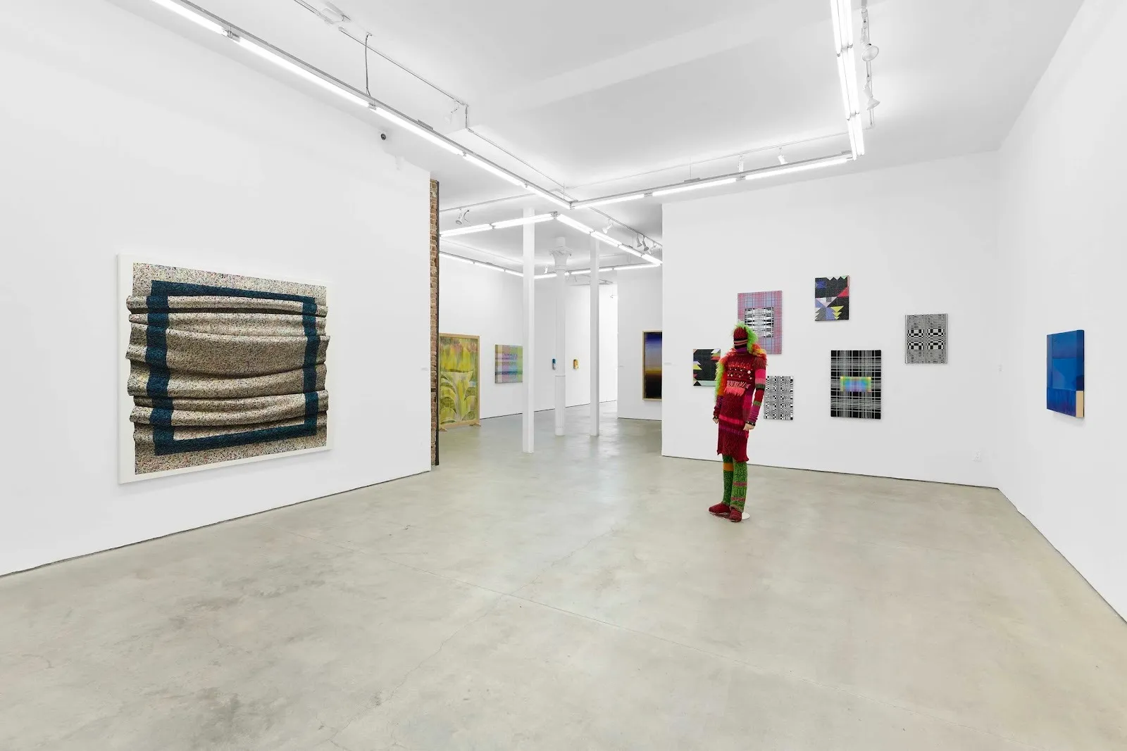 A white gallery complex features multiple textile and wall works of various sizes and colors. In the floor space, a colorful fabric mannequin, dressed in ornate designs, stands.