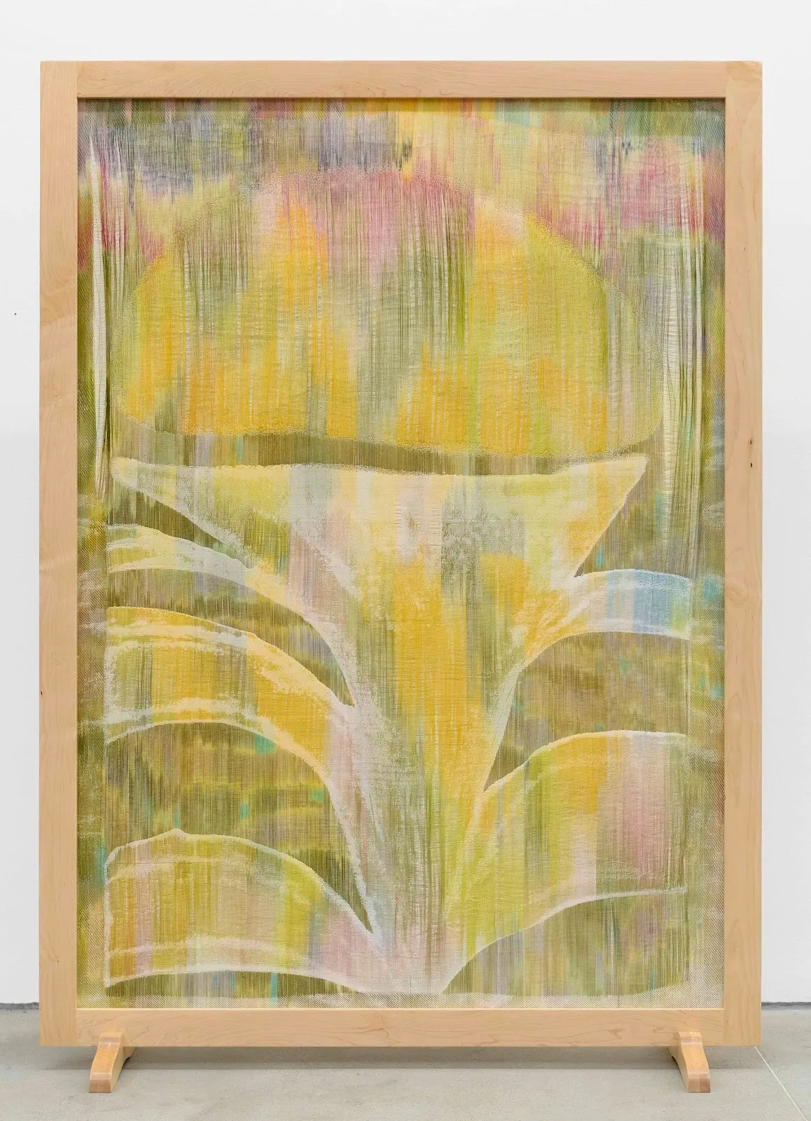 A large wooden frame contains a fiber work featuring watercolor-like washes of yellow, pink, blue, and green. An abstract, plant-like shape makes up the majority of the work.