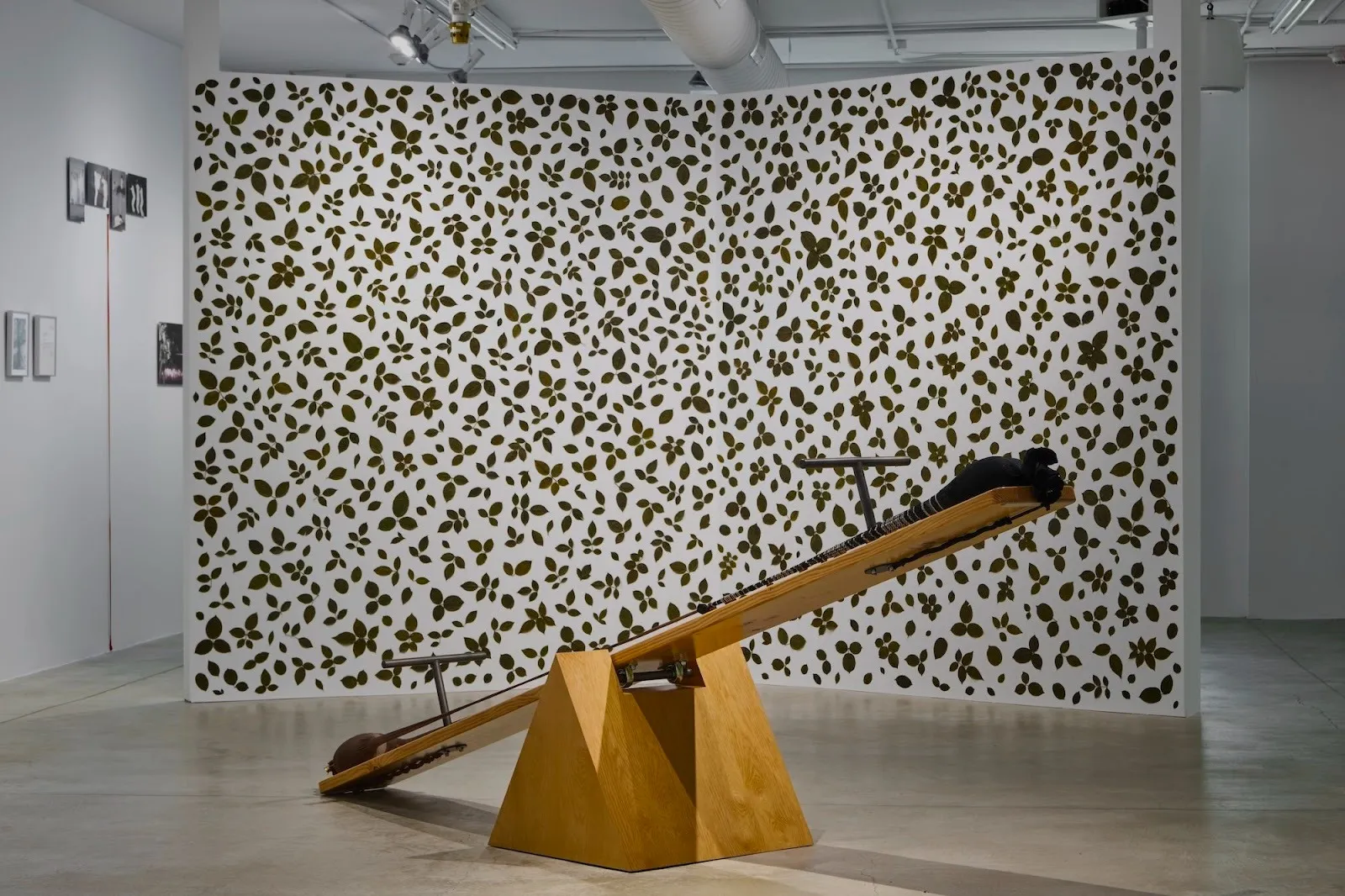A wooden seesaw-like sculpture with metal supports positioned in front of a large wall covered in a repeating olive-green leaf pattern.