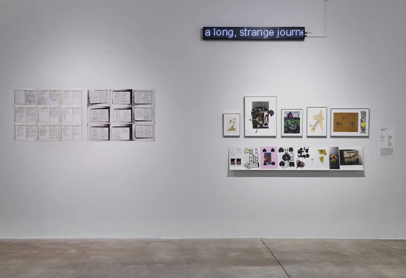 A white gallery wall displaying handwritten text panels, small framed mixed-media works on a shelf, and an LED sign reading “a long, strange journ—”.