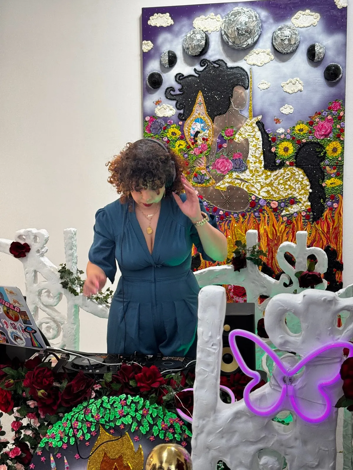 Amidst colorful, textured 2D works and sculptures, a person in a blue jumpsuit leans over a DJ set.