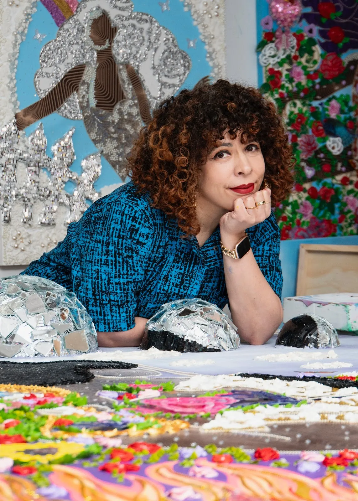 A person rests their chin on their hand and smiles directly at the camera. They lean on and are surrounded by colorful works of art.