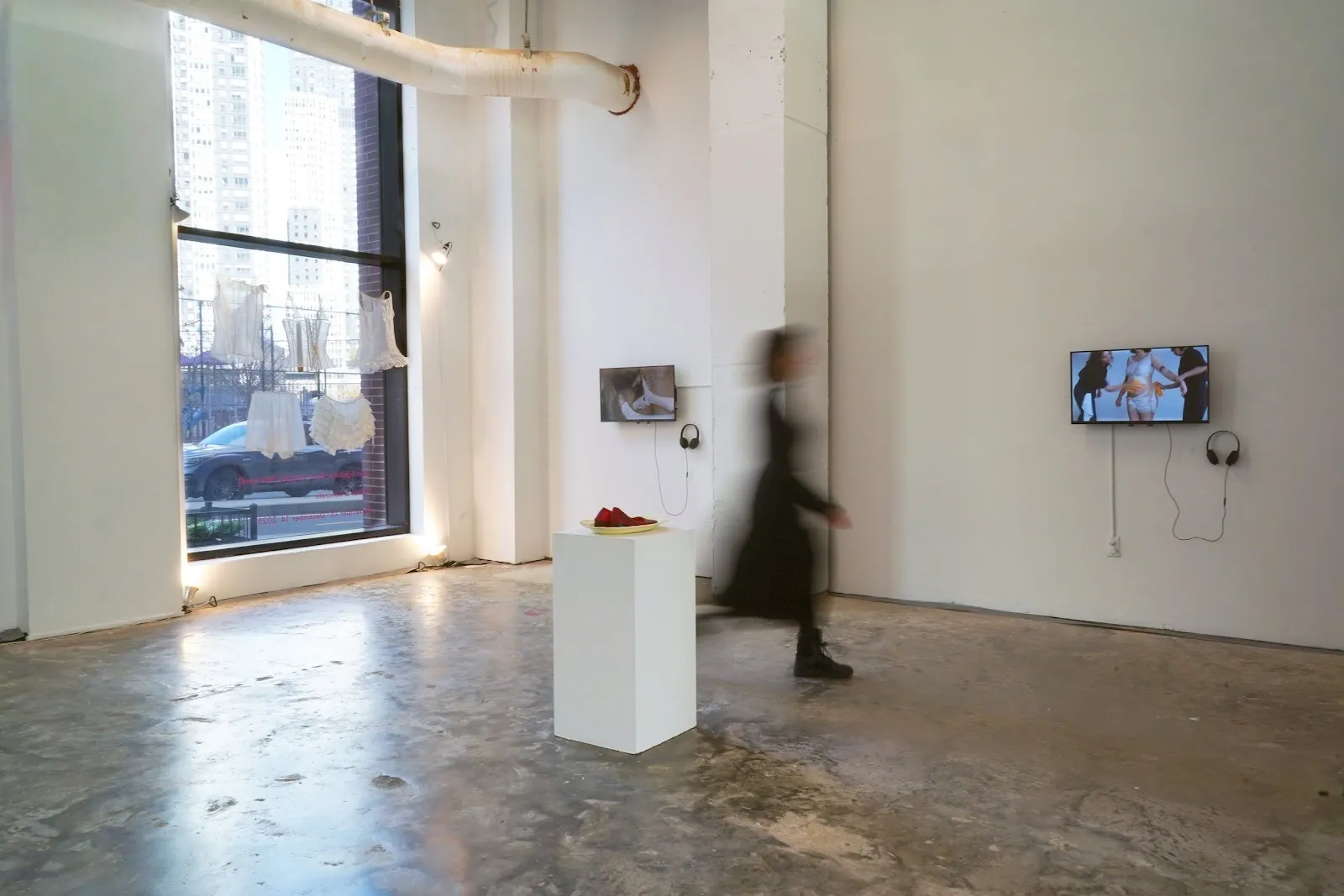 A white gallery space features wall-mounted monitors with headphones displaying different performance works. A squat square plinth supports an indiscernible dark red sculpture.
