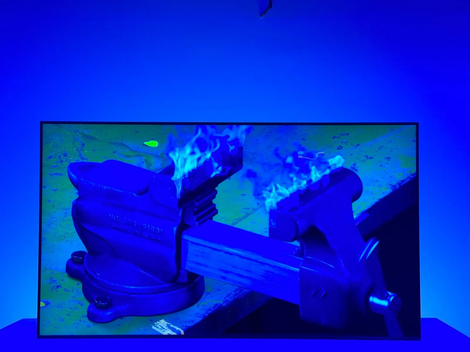 Room in blue neon light with tv monitor screening a video of anvil burning in flame, Gregory Kalliche, Anvil (2025). 4K video with sound, 9-channel lighting program, DMX controllers, lighting interface.
