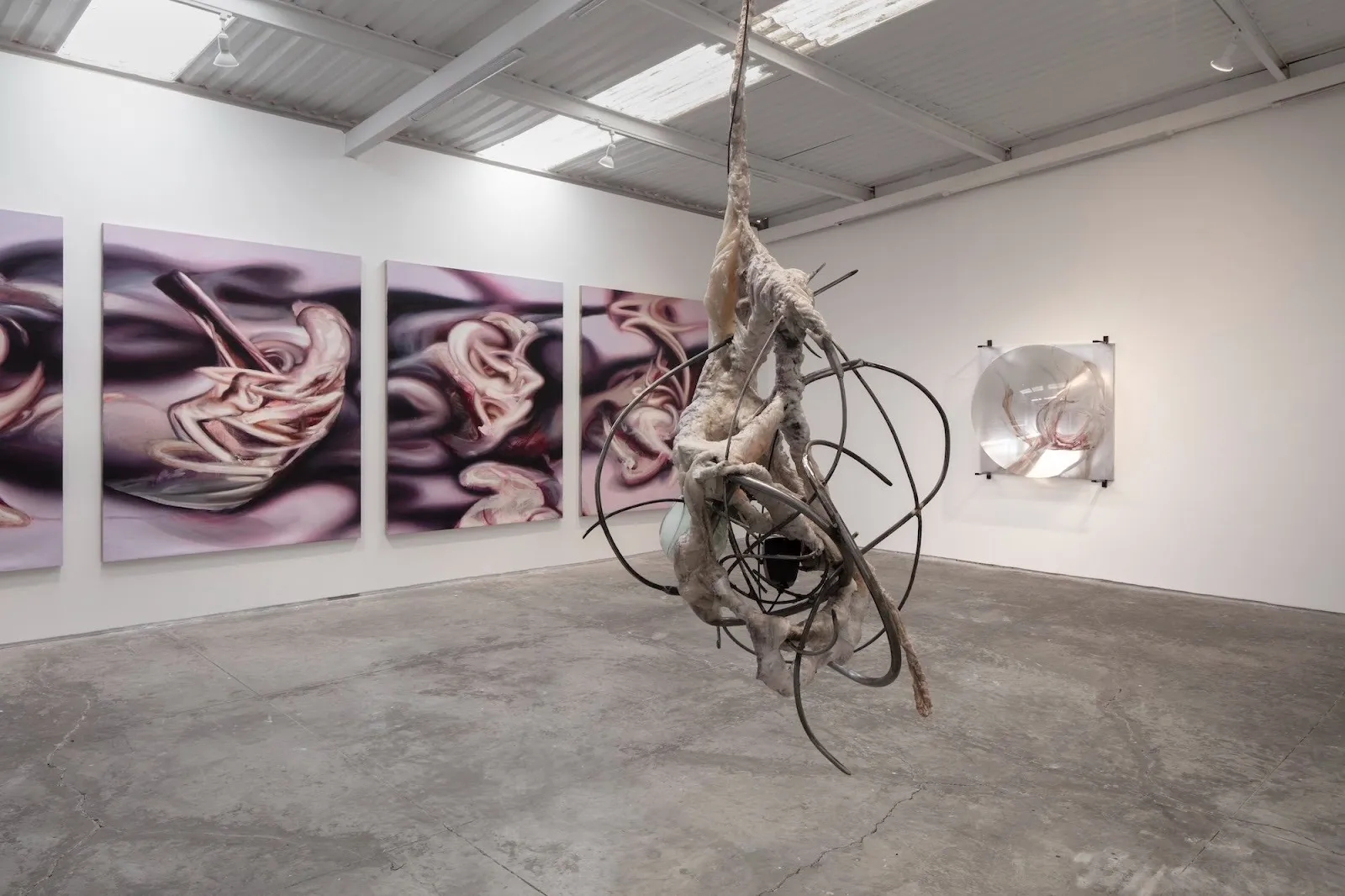 Hanging sculpture composed of pale, textured organic matter entwined with curved metal rods, shown against abstract paintings.