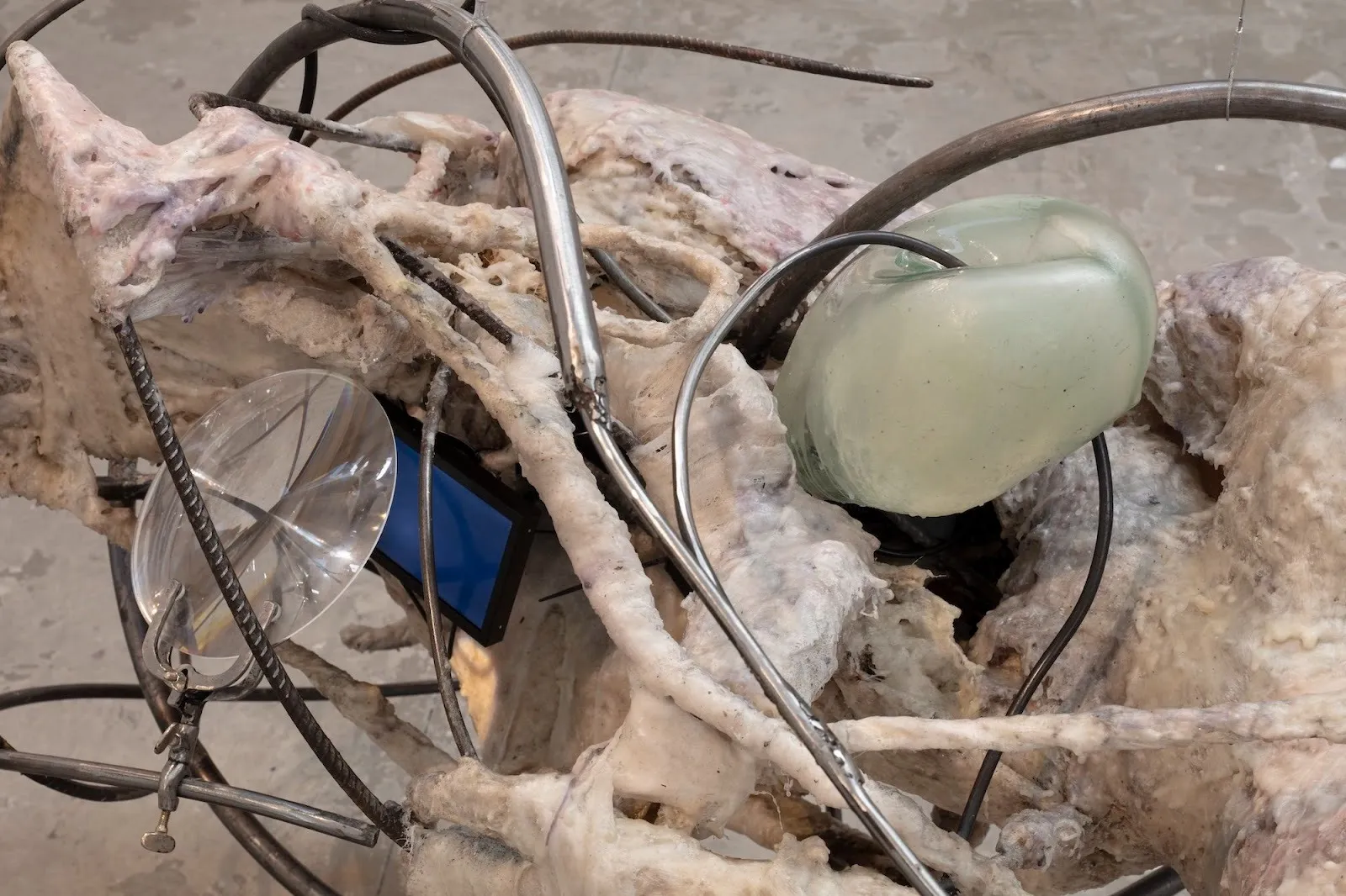 Close-up of a sculpture combining fibrous, skin-like material, bent metal tubing, translucent glass elements, and embedded electronic components.