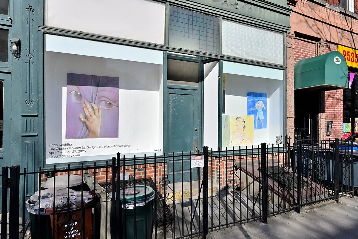 Side view of a store windowfront with paintings featured. The storefront is bordered with iron gates and trash cans are visible.