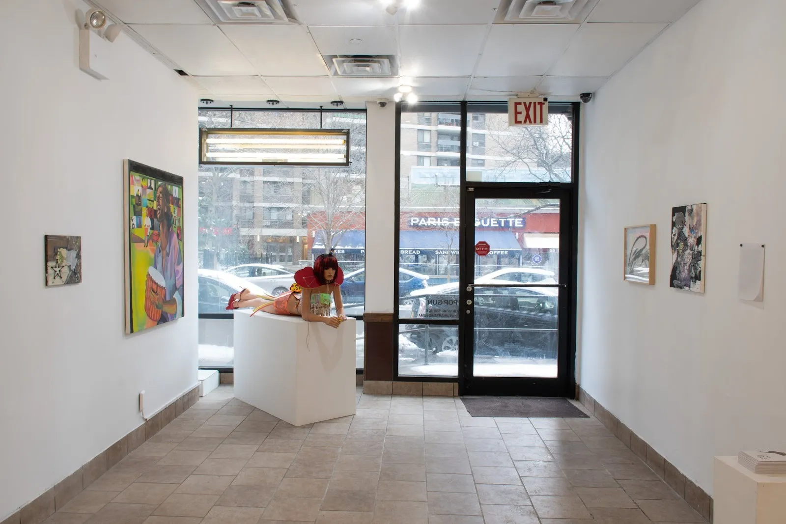 A storefront property houses both 2D and 3D works of art. A sculpture of a mannequin longing on a plinth stands next to the entryway. Outside, the street and buildings can be seen.