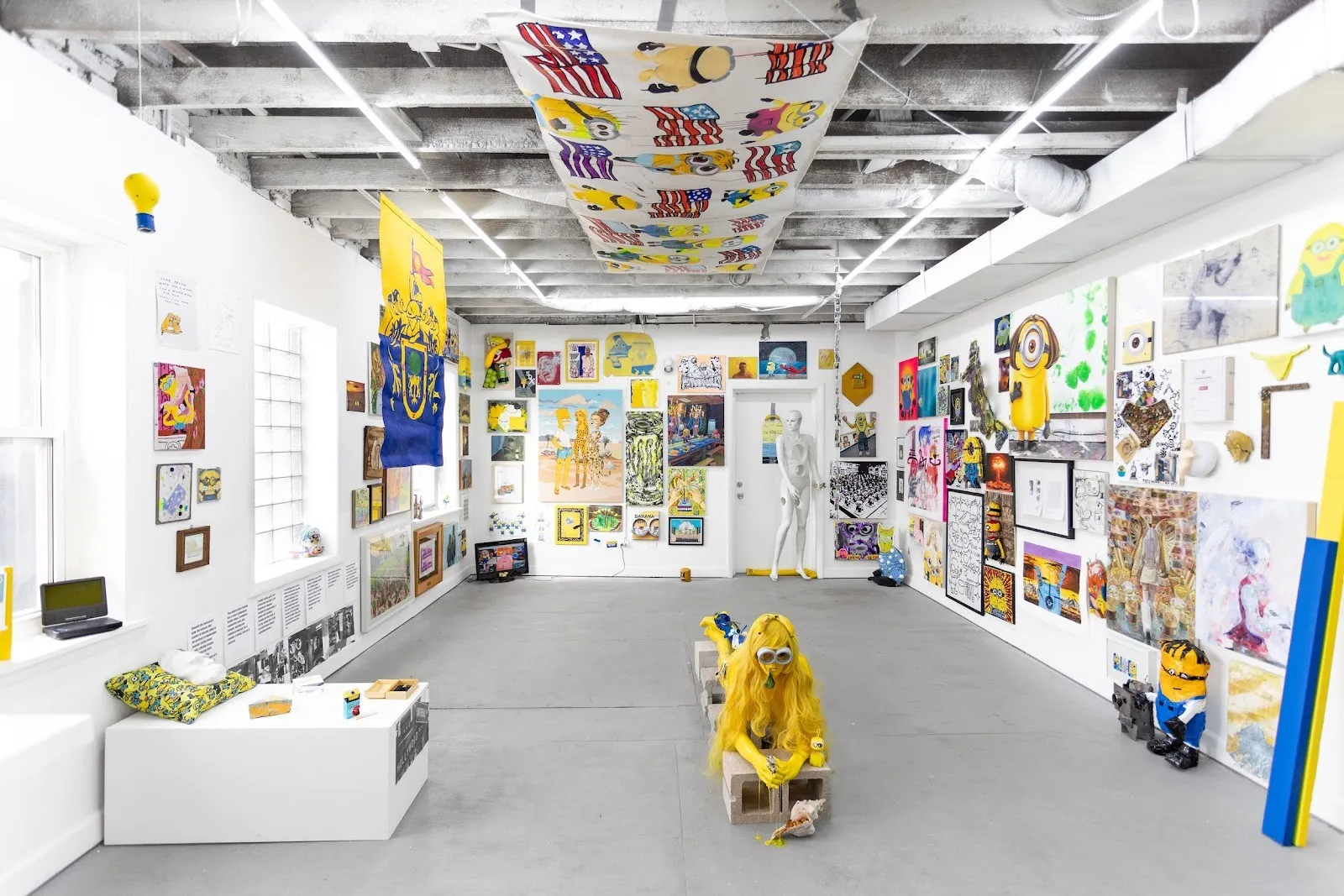A single-room gallery space is densly covered with works of 2D, 3D, and hanging art. Many of the works feature yellow and reference the minions from the "Despicable Me" movie franchise.