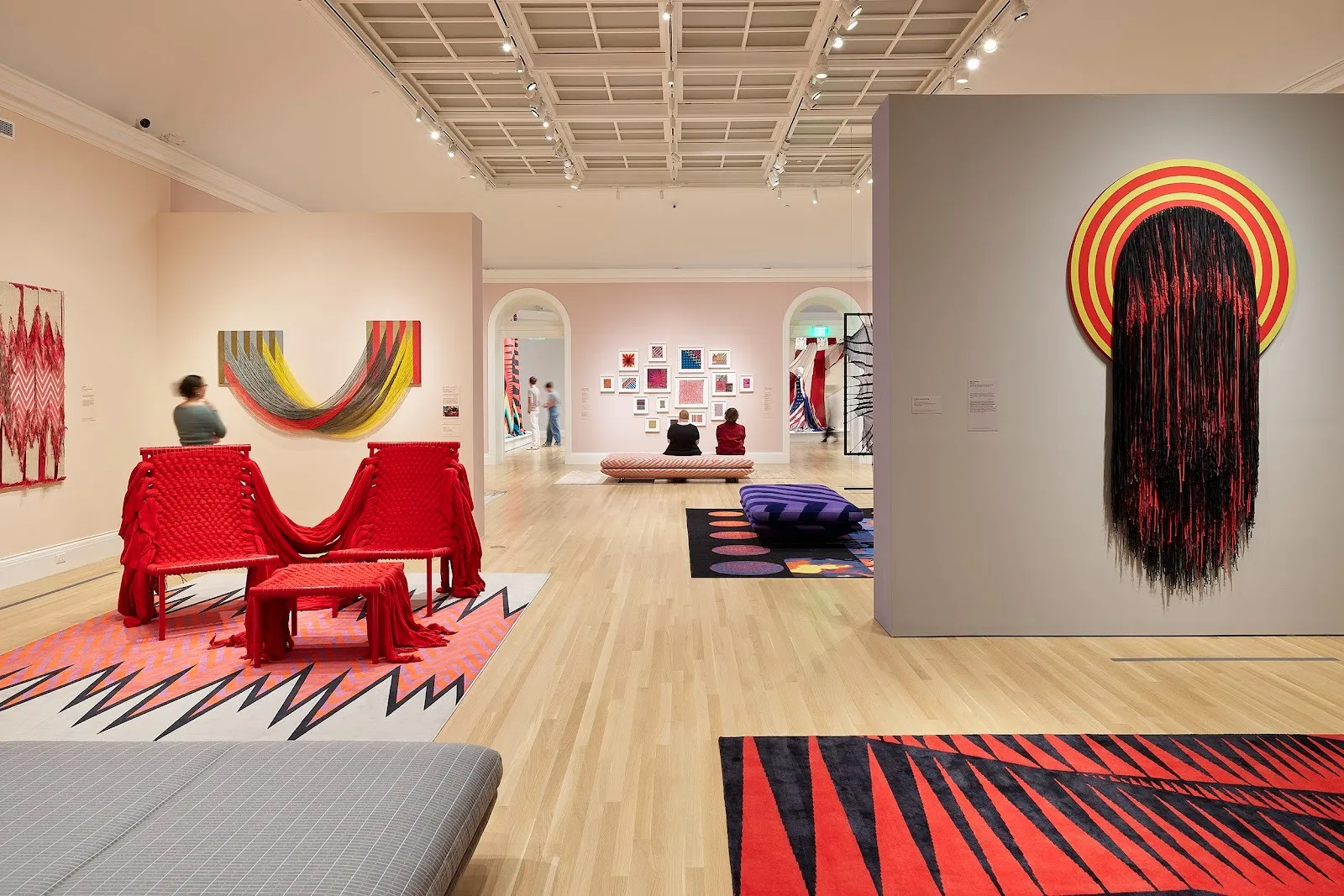 A large gallery room with light pink and grey walls feature a number of wall works and sculptural pieces. All works are made of textile, from woven patterns to draped threads. One sculptural piece features red chairs woven together.