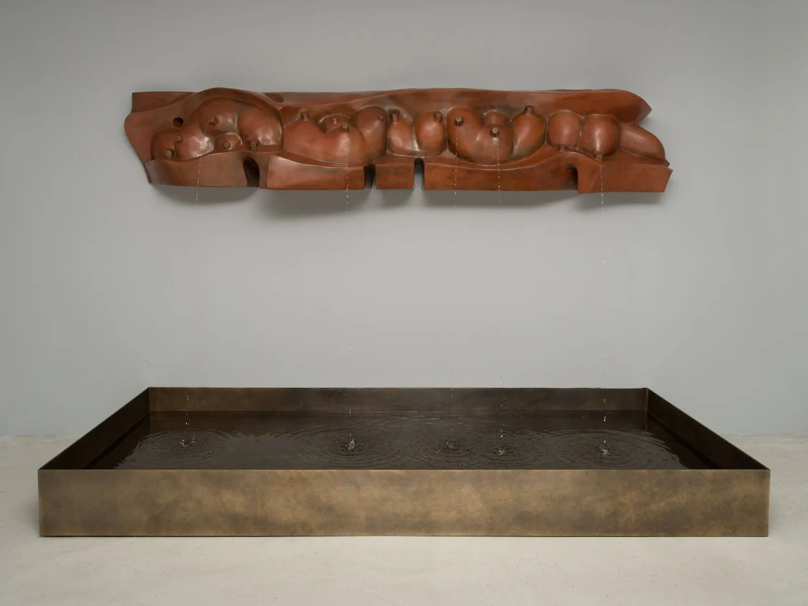 In a white gallery room, a low rectangular metal trough holds a pool of water. A sculpture mounted above to the wall made of metal cast in organic, bulbous forms drips streams of watter from nipple-like openings into the trough.