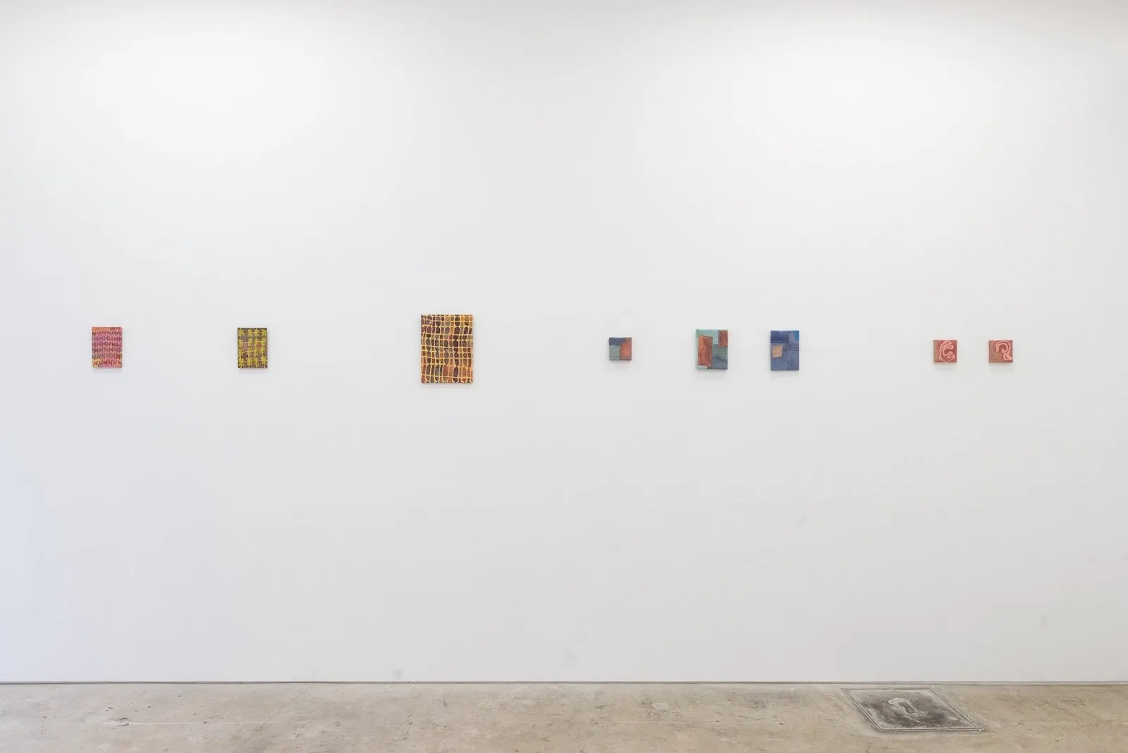 A white gallery wall features a number of small, rectangular works. Each features soft colors and cross-weave patterns.