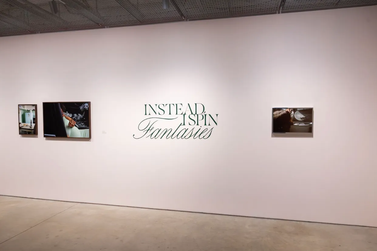 A white wall features three colored photographs sandwiching wall text that reads "INSTEAD, I SPIN FANTASIES" in green.
