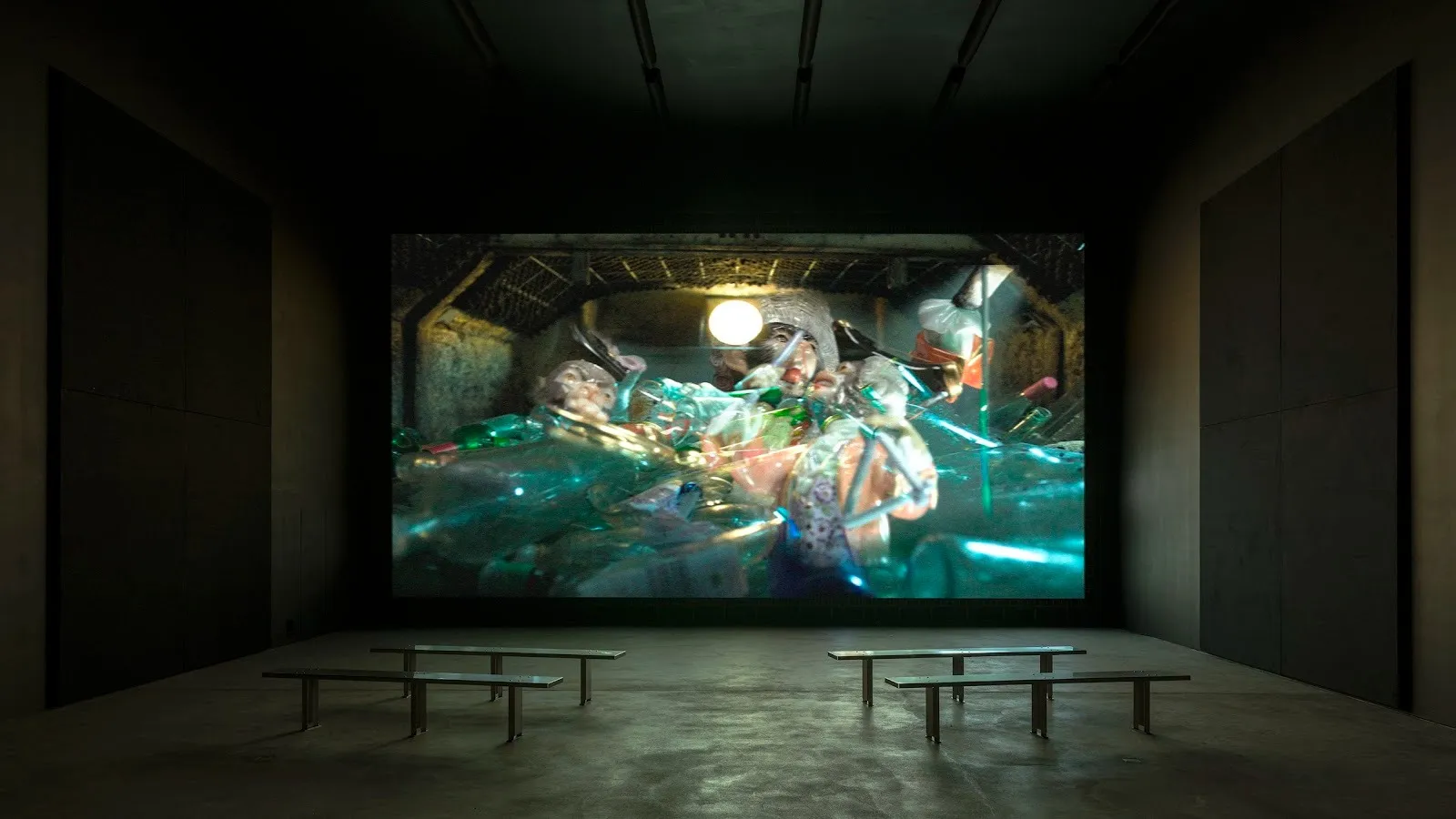 A plain, darkened room with four benches angled toward a large, floor-to-ceiling screen. The screen depicts an ambiguous shot of iridescent folds of cellophane in a sort of lit box.