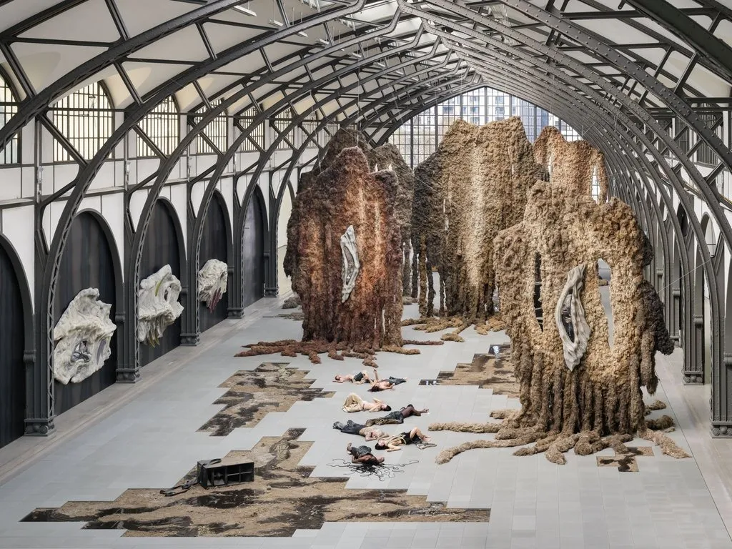A large, warehouse-hall-hangar-like building houses massive hanging and 3D works, as well as performance artists who lie on the floor. The large, hanging works seem to be made of fabric-like, tan and brown tendrilous masses.