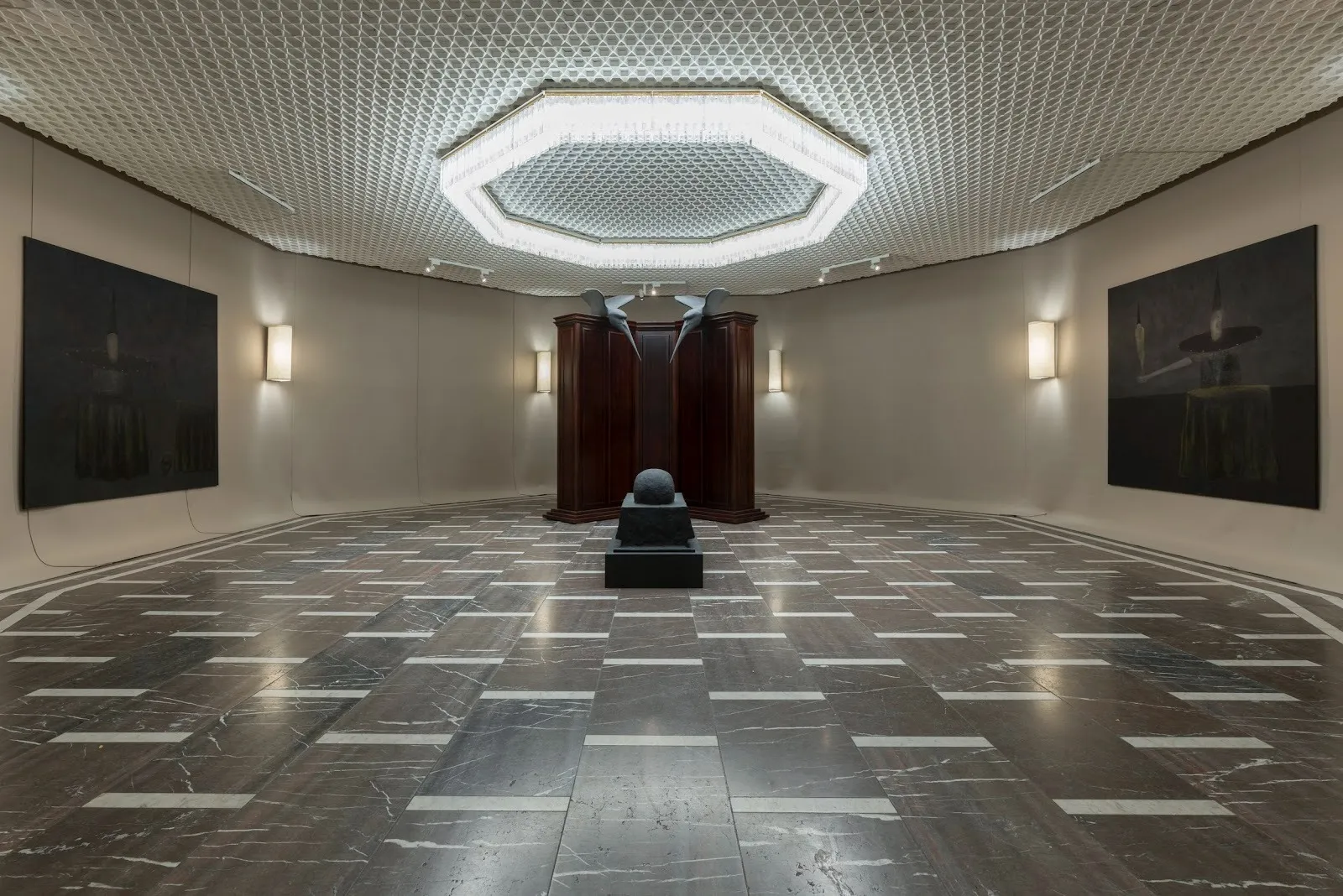 An octagonal room features an octagonal chandelier, black and white marble floors, and wall sconces. 2D works hang on the wall and a central stone sculpture sits at the center of the room, with two more gargoyle-like sculptures hanging from a doorway
