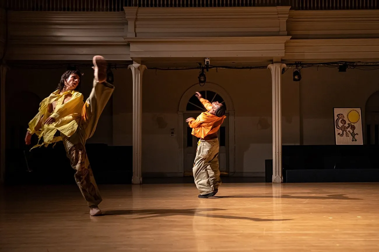 On a spotlit stage space with a balcony in the background, two dancers are caught mid-movement. In orange and yellow clothes, they spin and throw their arms and legs up, heads thrown back or looking upwards.