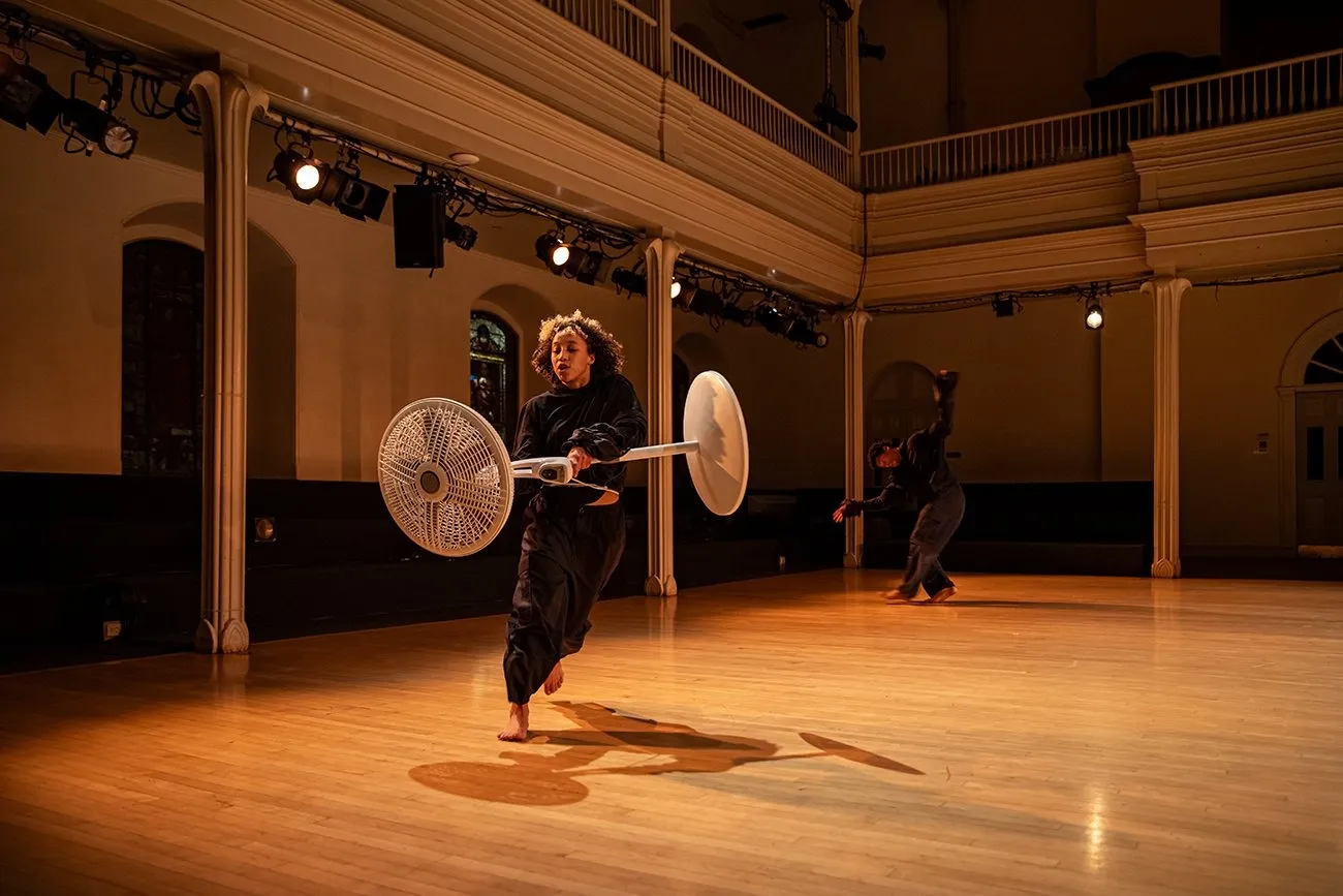 In a balconied performance space, a dancer carries a white standing fan and runs about the space. In the background, another performer gestures with outstretched hands and bent legs.