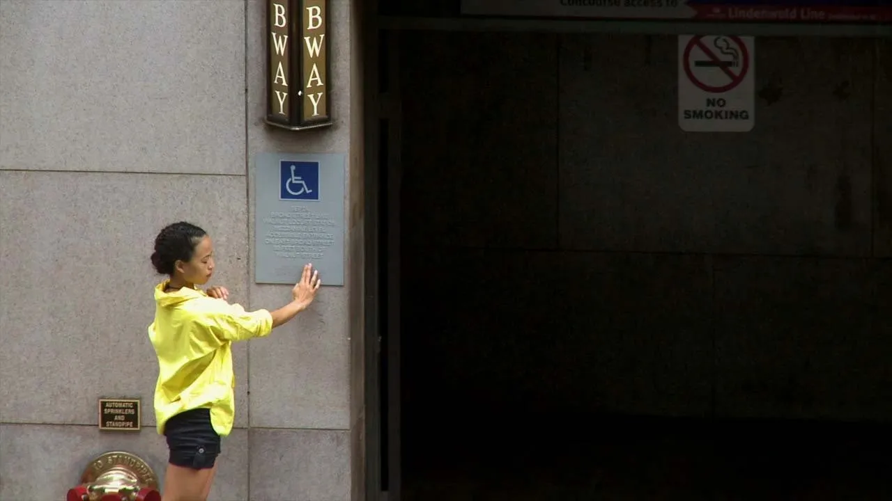 Outside a building with a double-sided light-up sign that reads "BWAY," a person in a yellow button down shirt fingers a handicapped braille sign.