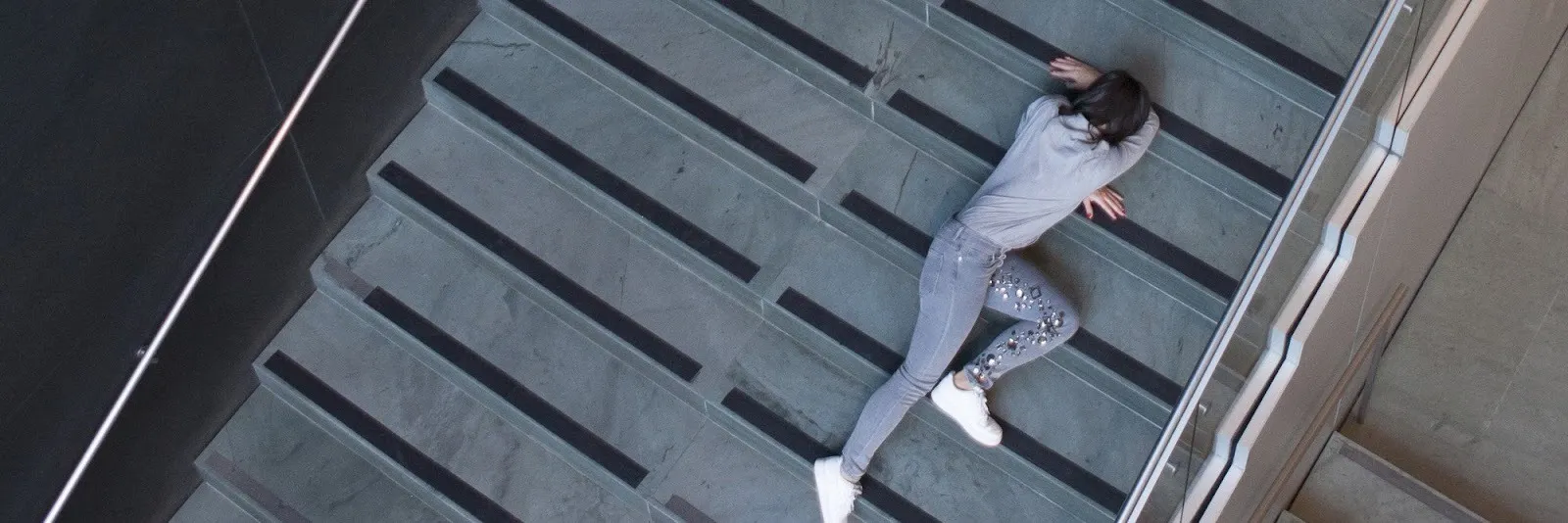 An aerial shot of a grey stone staircase with a person clad in grey sprawled across it, their face cradled in one of their arms resting on the steps.