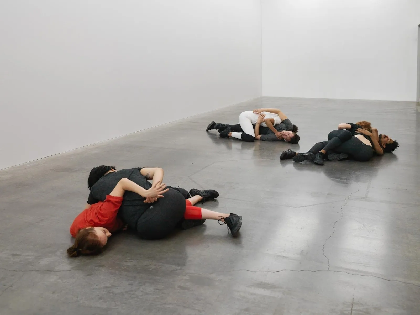 On a concrete floor in a white-walled space, performers clad in black, white, and red drape and embrace each other in contorted positions—half choreography, half wrestling-like.