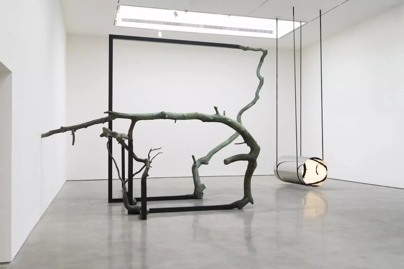 A two-part sculpture, or two separate sculptures, sit in a gallery room. One is made from large, rectangular metal bars which morph into tree branches, the other is a low-hung metal tube, illuminated from inside.
