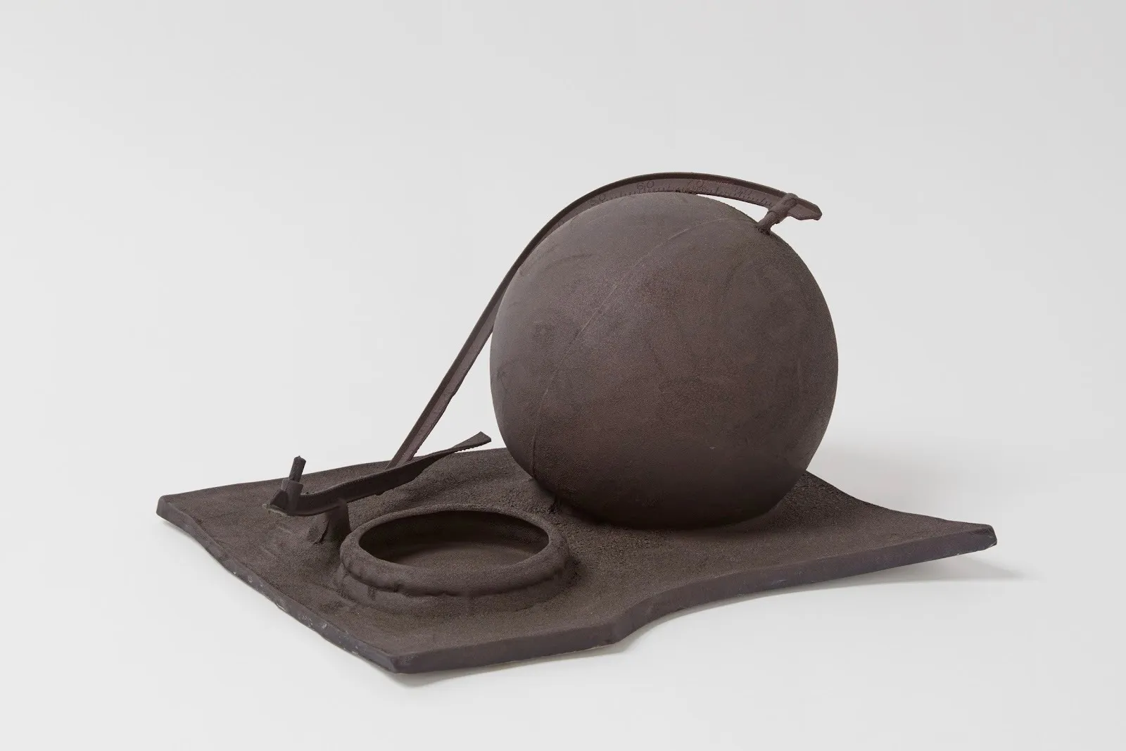 Dark, matte sculptural assembly with a sphere and bent rods resting on a flat, uneven base.