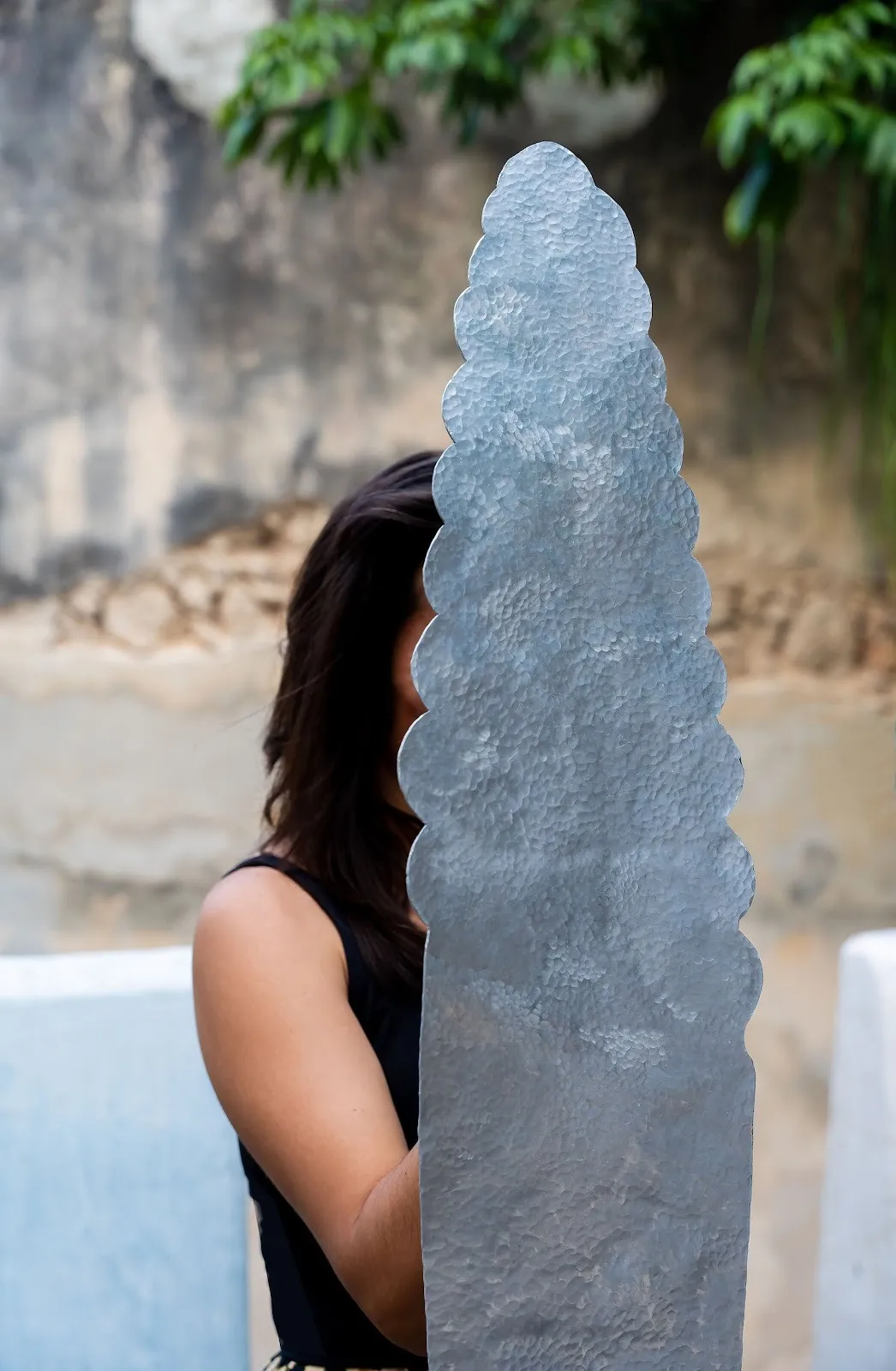 A person partially obscured behind a tall, scalloped silver metal form held upright outdoors.
