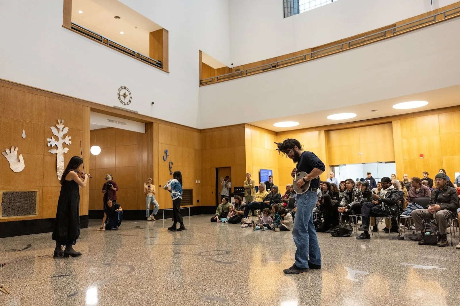 Performance in a wood-paneled atrium where musicians and dancers move among silver metal wall pieces as an audience watches.
