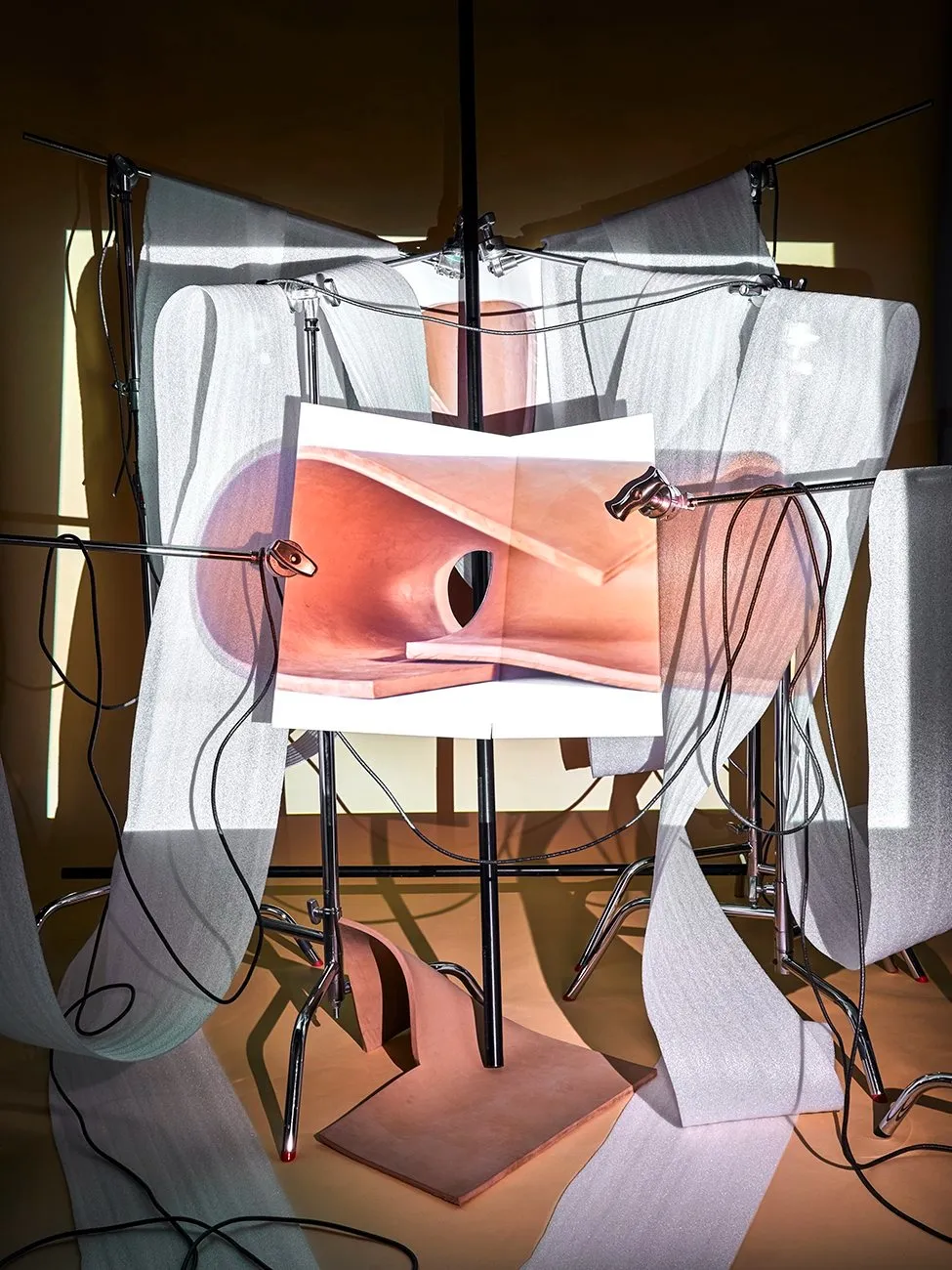 Intricate studio setup with lighting equipment, draped white fabric, cables, and a central photograph of a curved, flesh-toned architectural form.