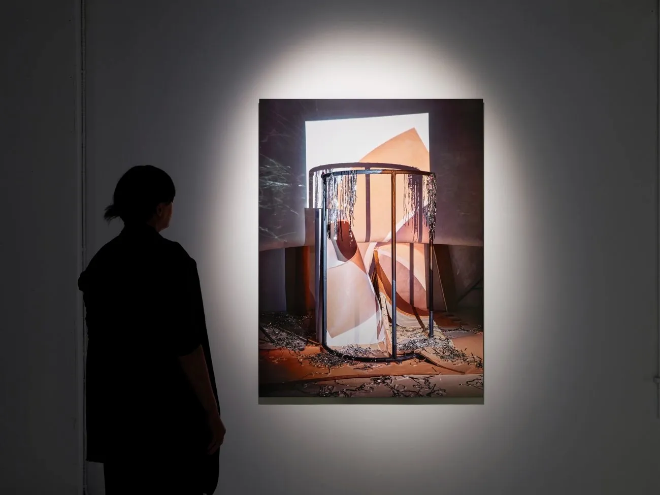 A person views a large, spotlit photograph depicting a cylindrical metal cage and abstract forms, with metal fragments on the floor.