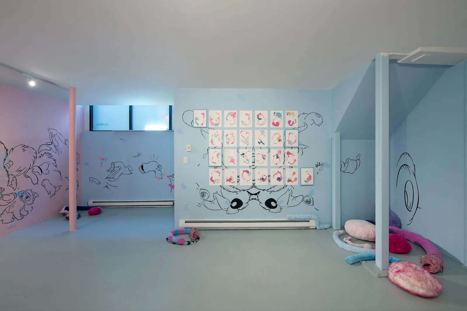 A gallery room's walls are painted light pink and blue. Along the walls, cartoonish line drawings are painted on. Colorful, plush sculptures are scattered in the corners of the space. A grid of works on paper take up much of the central wall.