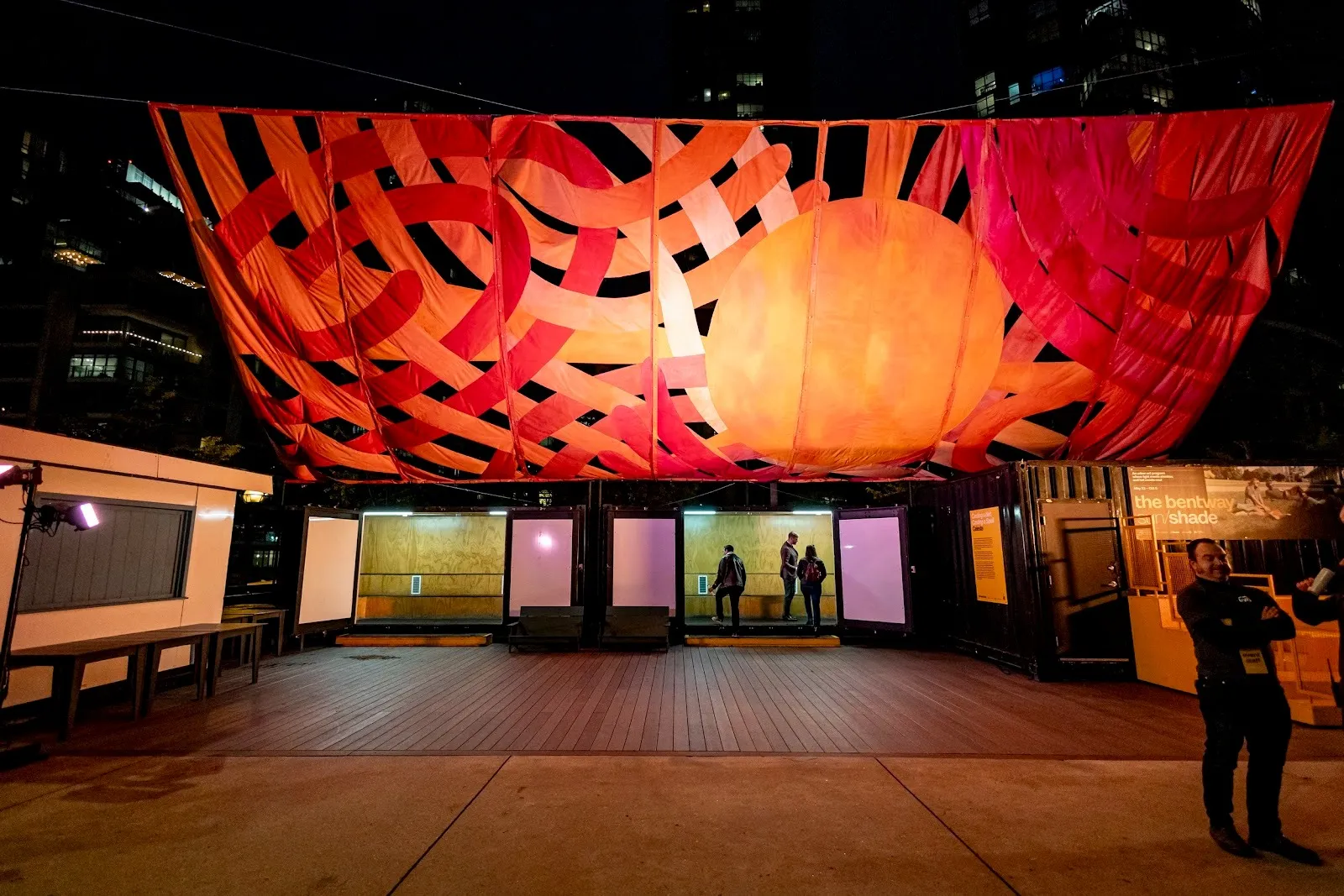 Over an outdoor space at night, a large, canopy-like textile work stretches over. The canopy is made from strips of curling red and orange fabric crossing over itself..