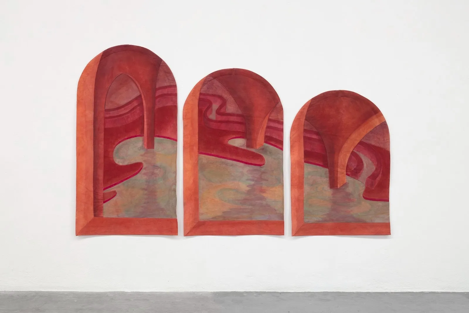 A 2-D wall piece is made of three distinct parts positioned closely in a row: arched structures that decrease in height. Together, the pieces form an architectural landscape made of reds and light blue.